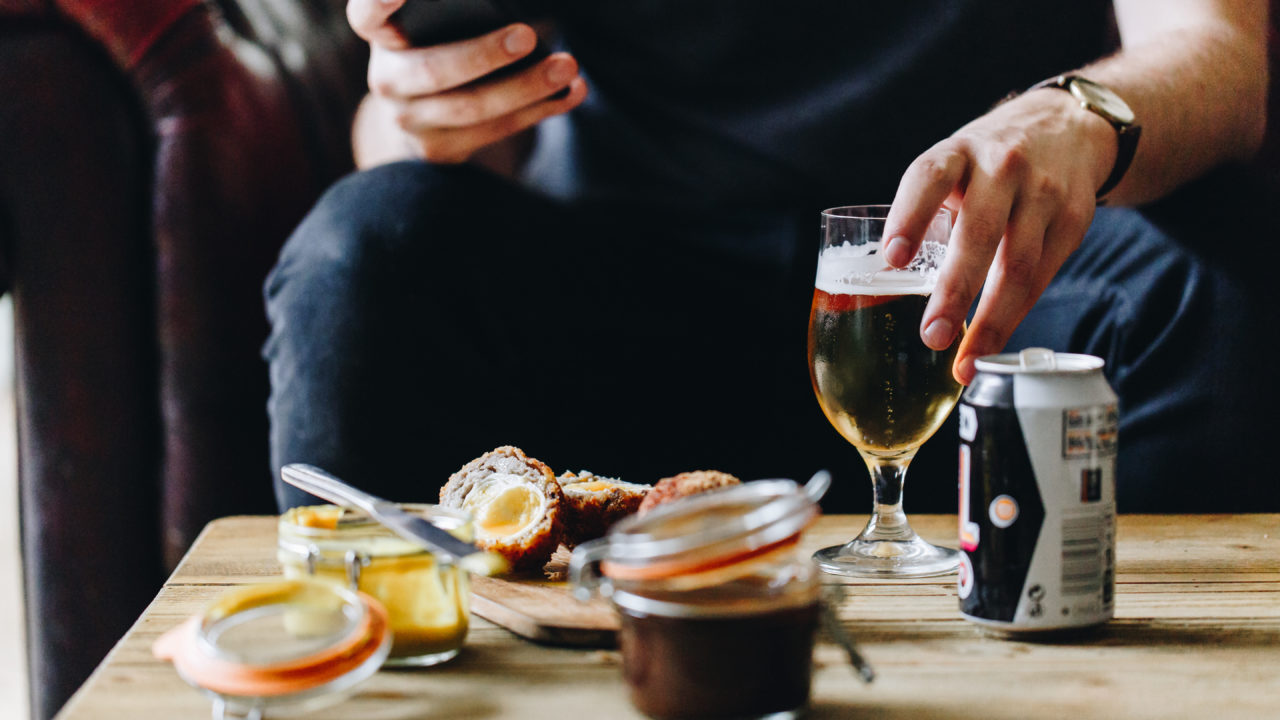 Person drinking sitting on a sofa with a drink and scotch egg on the table in front of them