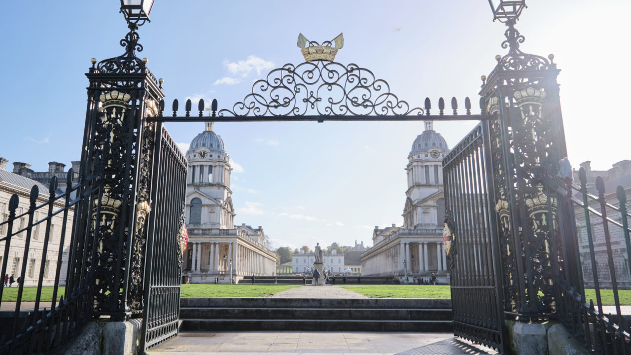 A grand entrance gate with intricate black and gold detailing opens up to a view of the Old Royal Naval College in Greenwich, London. The symmetrical architecture features two domed buildings flanking a central walkway, leading to a statue and further buildings in the background. The sky is clear with the sun shining brightly, casting light over the entire scene.