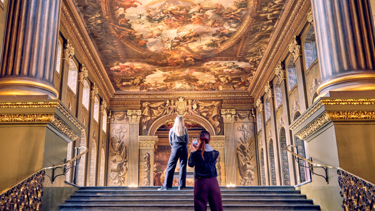 Two people stand at the top of a grand staircase inside the Painted Hall at the Old Royal Naval College in Greenwich, London. The hall is adorned with large columns and intricate gold detailing. The ceiling above them features a magnificent and elaborate mural filled with vibrant scenes and figures. One person is taking a photo of the mural while the other admires the artwork. The room is illuminated with warm lighting, highlighting the ornate decorations and grandeur of the space.